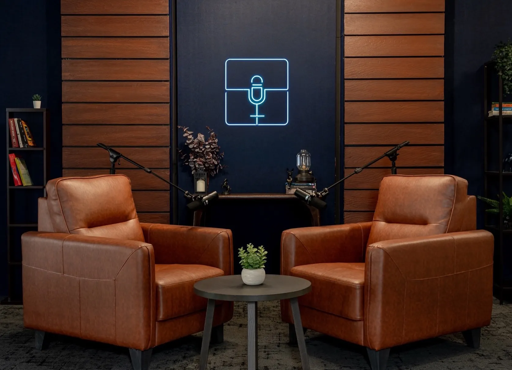 Two tan leather armchairs with boom-arm microphones in front of a navy wall with a blue neon microphone sign
